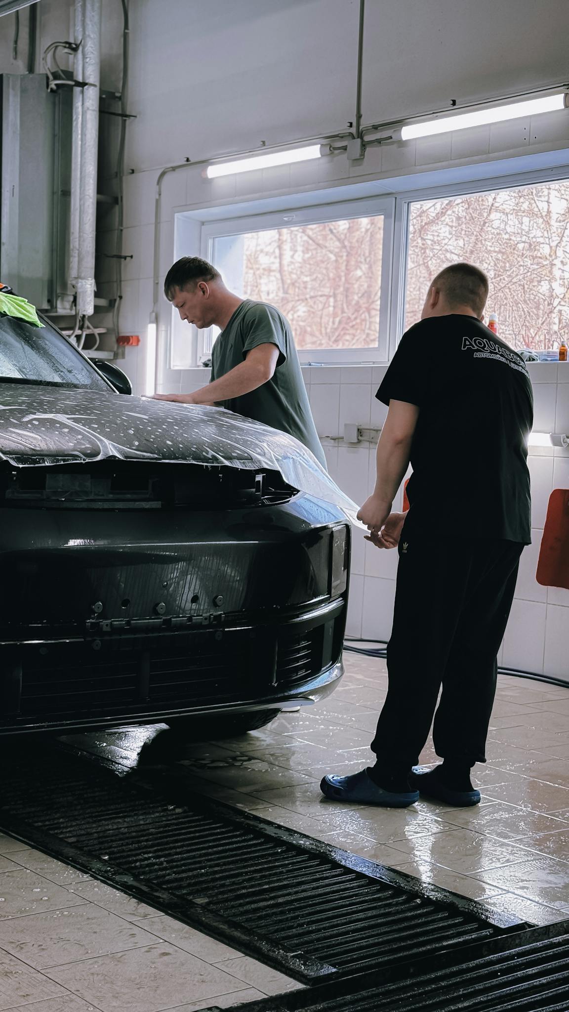 Two professionals applying protective film to a black car in an indoor workshop.